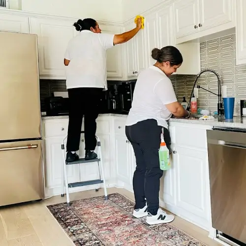 Two professional maids cleaning a bright white kitchen—one wiping upper cabinets on a step stool, one scrubbing the sink—house cleaning The Colony by Dallas Sunrise Maids in The Colony, TX