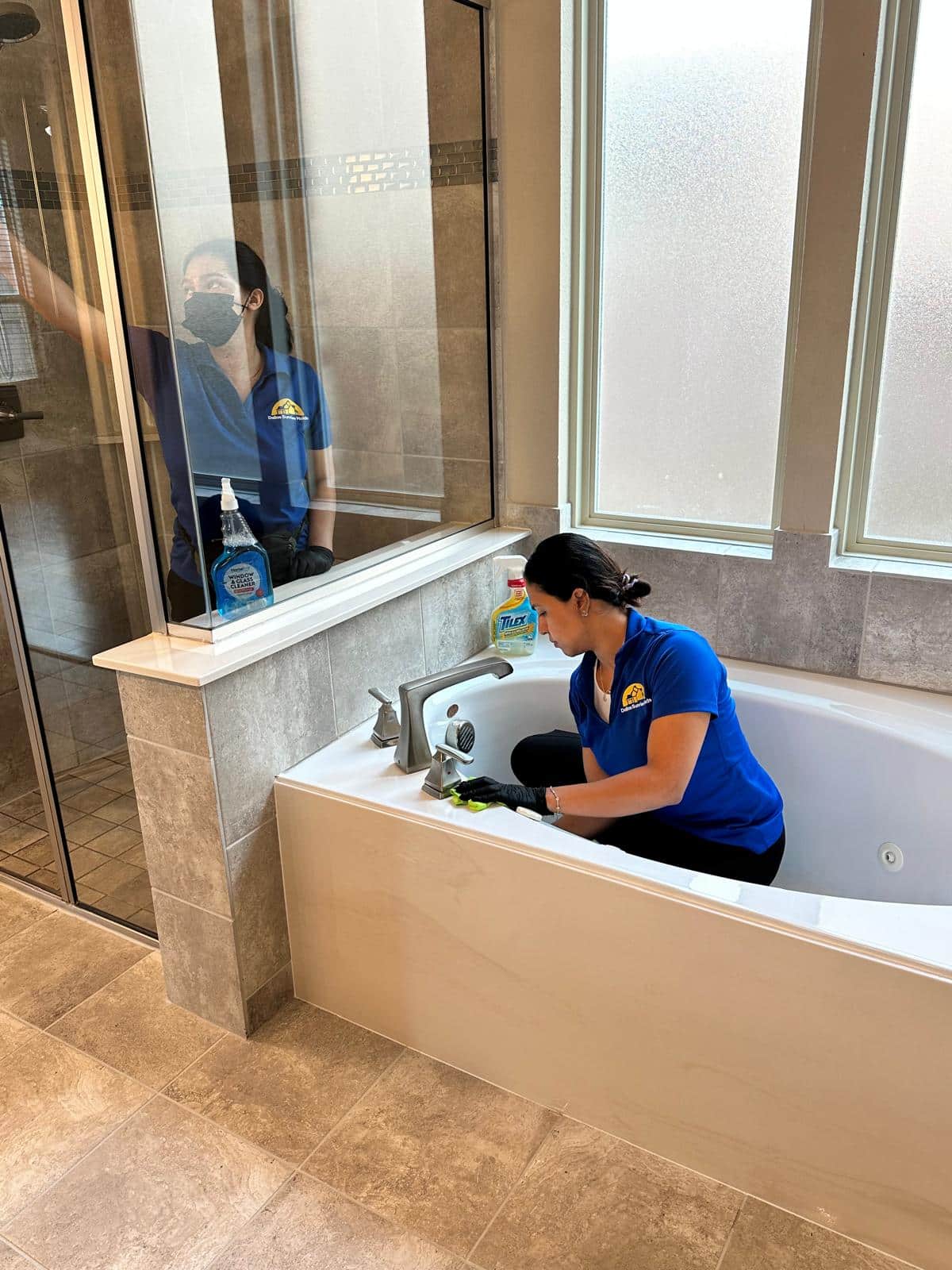 Two professional cleaners in blue uniforms scrubbing a large bathtub and wiping down glass shower doors, demonstrating how clean should my house be before a cleaner comes for home cleaning in Plano, Texas by Dallas Sunrise Maids.