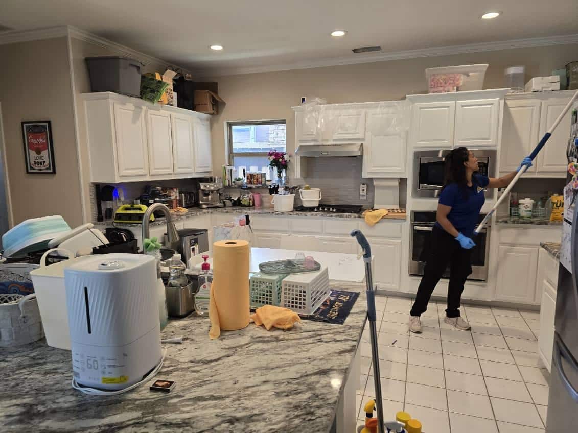 A professional cleaner in a blue uniform dusting high kitchen cabinets with an extension pole, answering the question of how clean should my house be before a cleaner comes for home cleaning in Plano, Texas by Dallas Sunrise Maids.