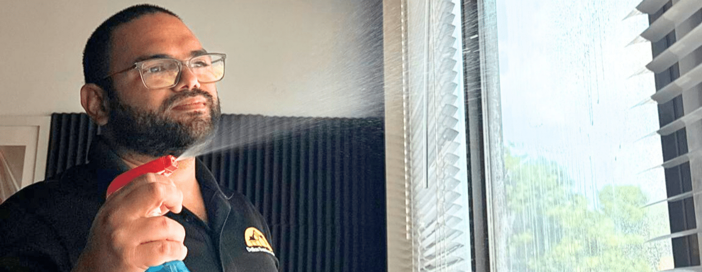 Jason Flores sunrise maids team member spraying a cleaning solution onto window glass with blinds raised, demonstrating home cleaning in Plano, Texas by Dallas Sunrise Maids.